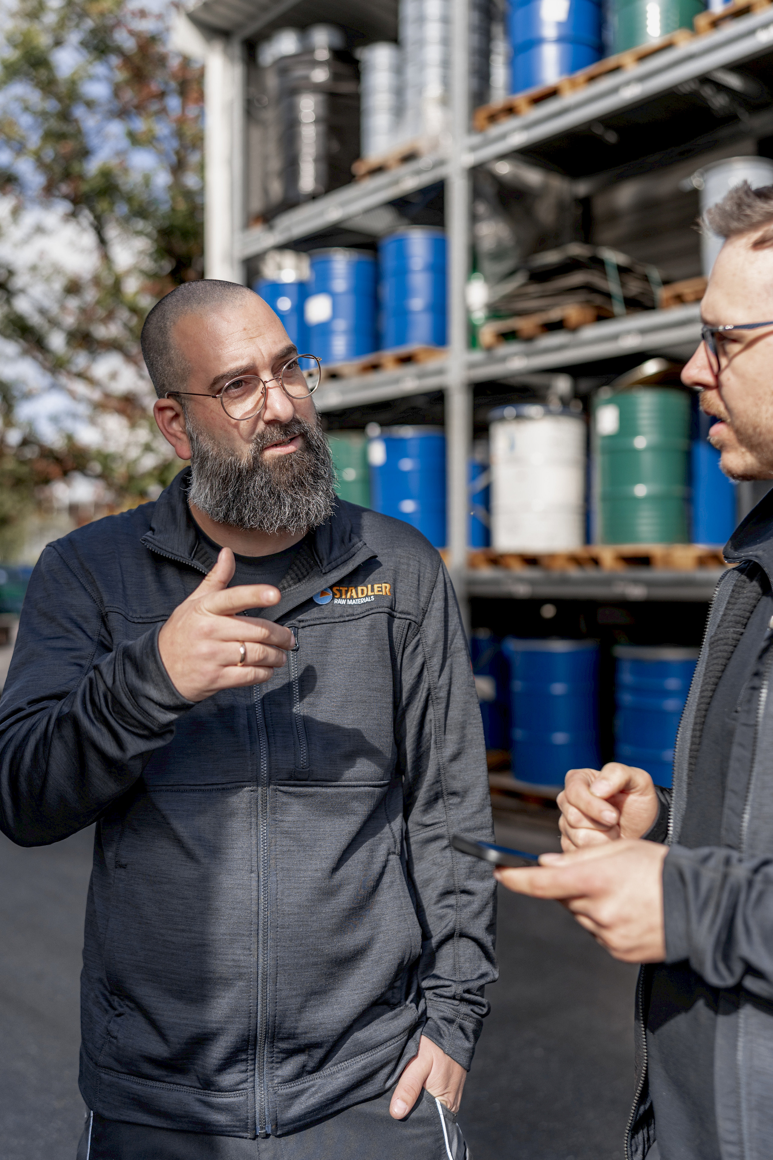 Zwei Männer diskutieren draußen vor einem Regal mit farbigen Fässern.