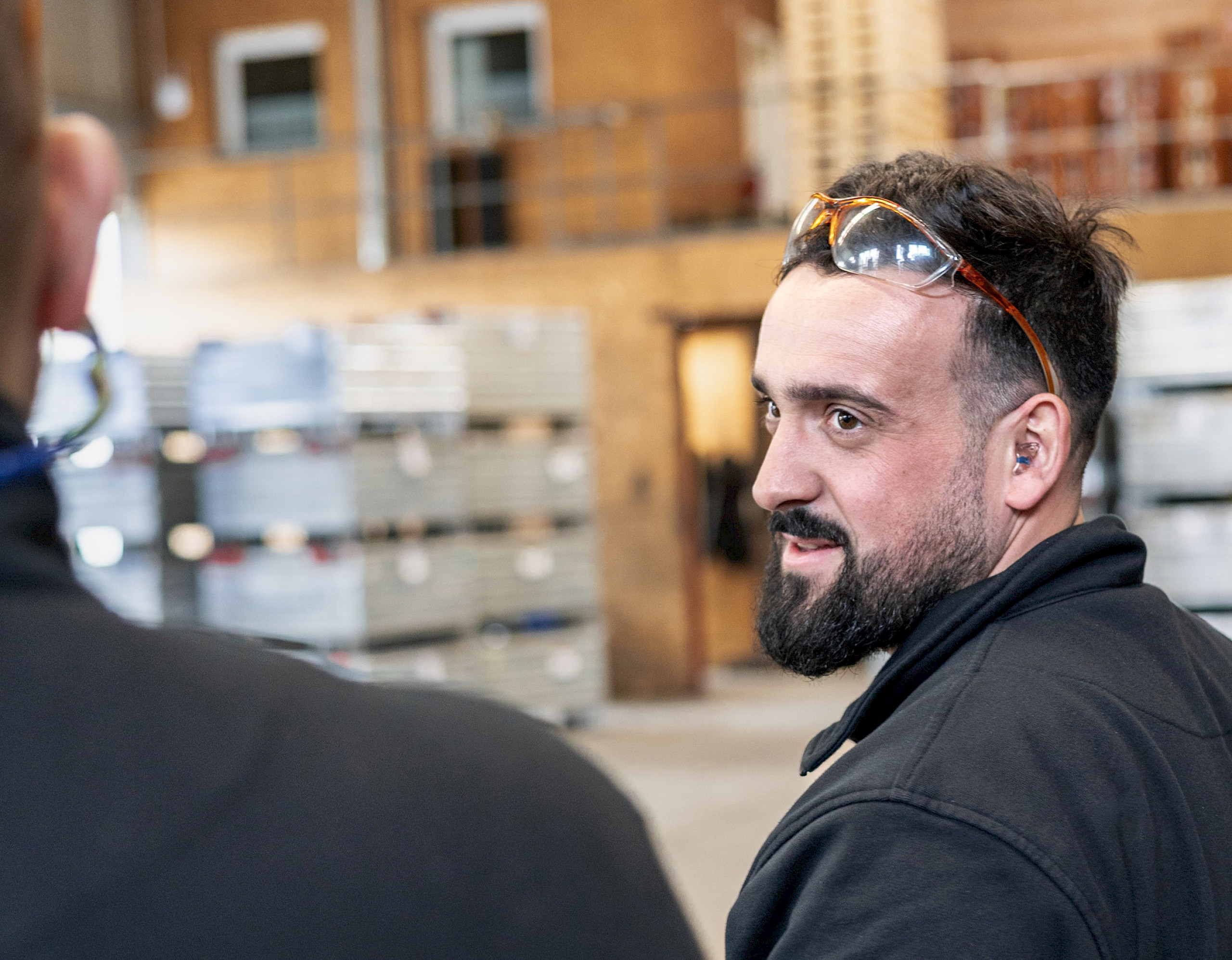 Ein Mann mit Sicherheitsbrille auf dem Kopf steht in einer Lagerhalle und schaut zur Seite. Im Hintergrund sind Regale mit Kisten zu sehen.