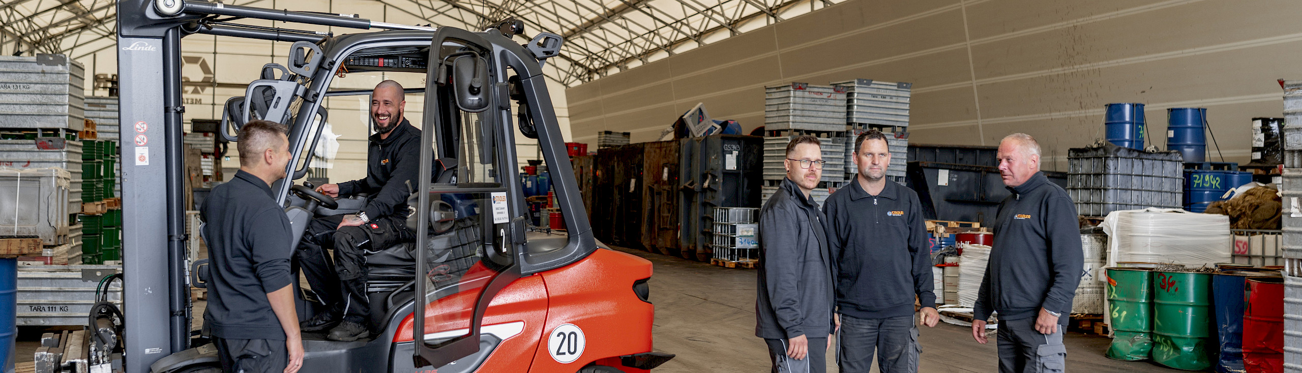 Fünf Männer in Arbeitskleidung in einer Lagerhalle, einer sitzt auf einem Gabelstapler, die anderen stehen um ihn herum. Im Hintergrund sind gestapelte Kisten und Fässer sichtbar.