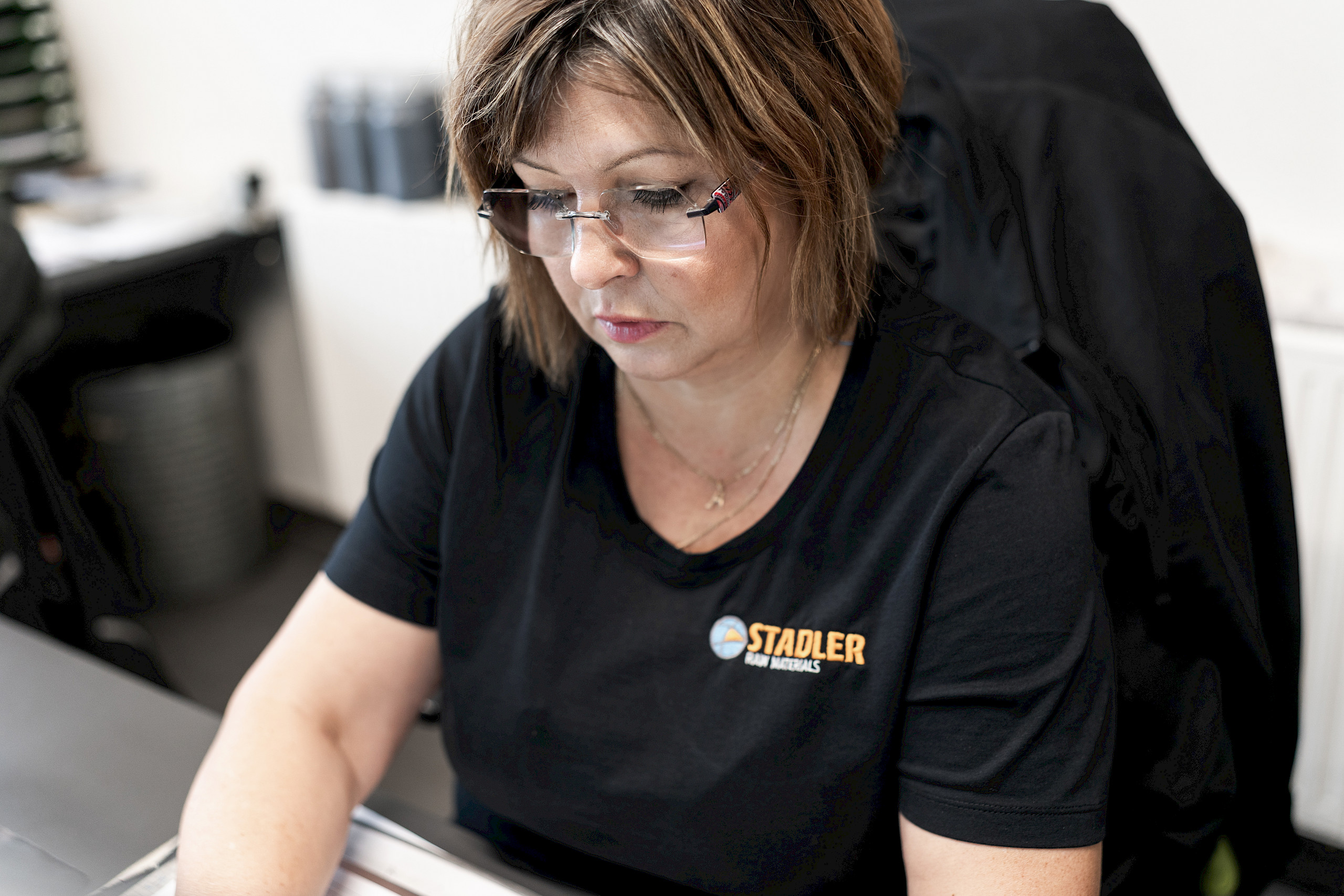 Eine Frau mit Brille arbeitet an einem Schreibtisch, trägt ein schwarzes T-Shirt mit einem Logo und einem Schriftzug.