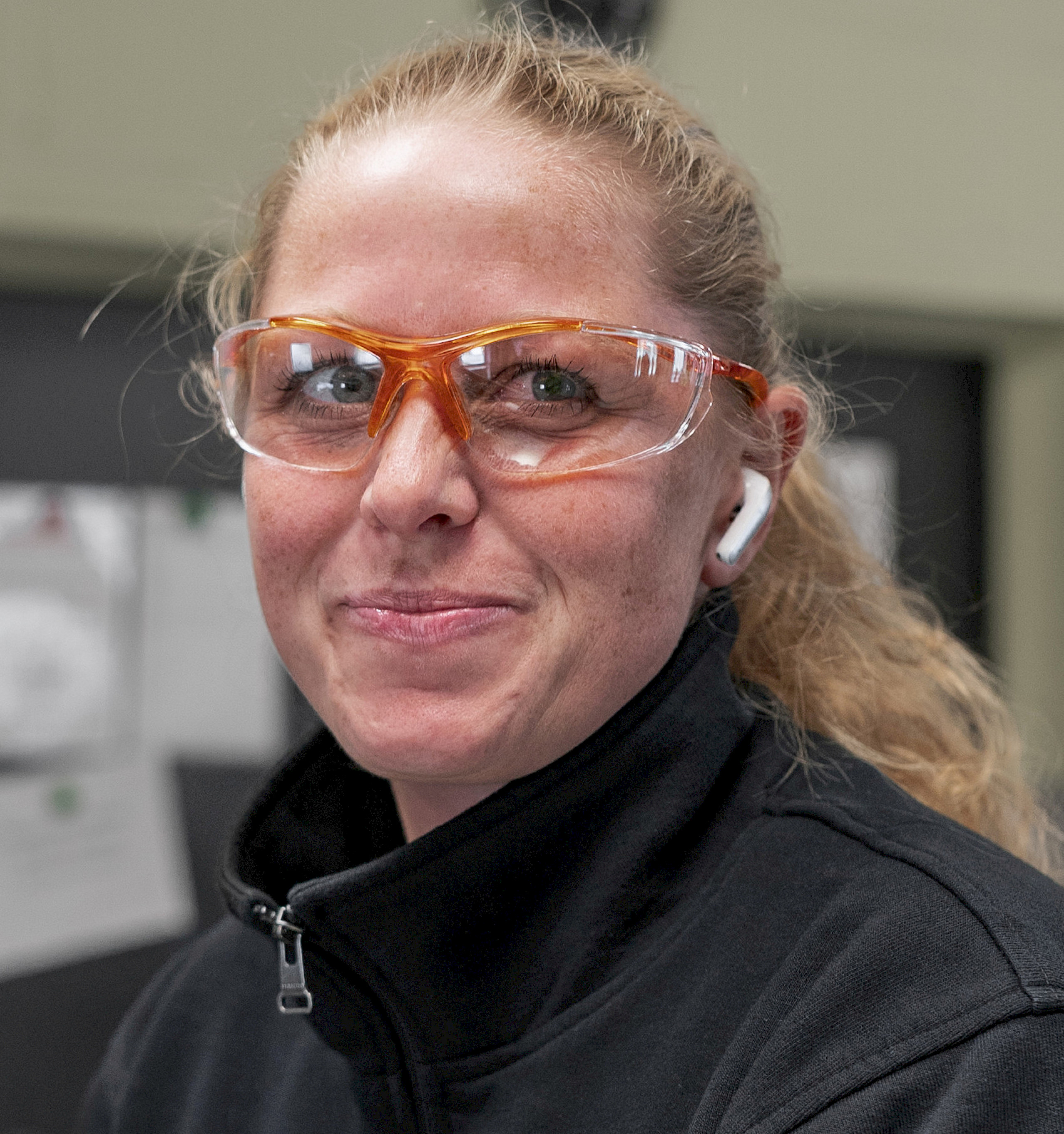 Eine Person mit Schutzbrille und Ohrhörern lächelt in die Kamera.