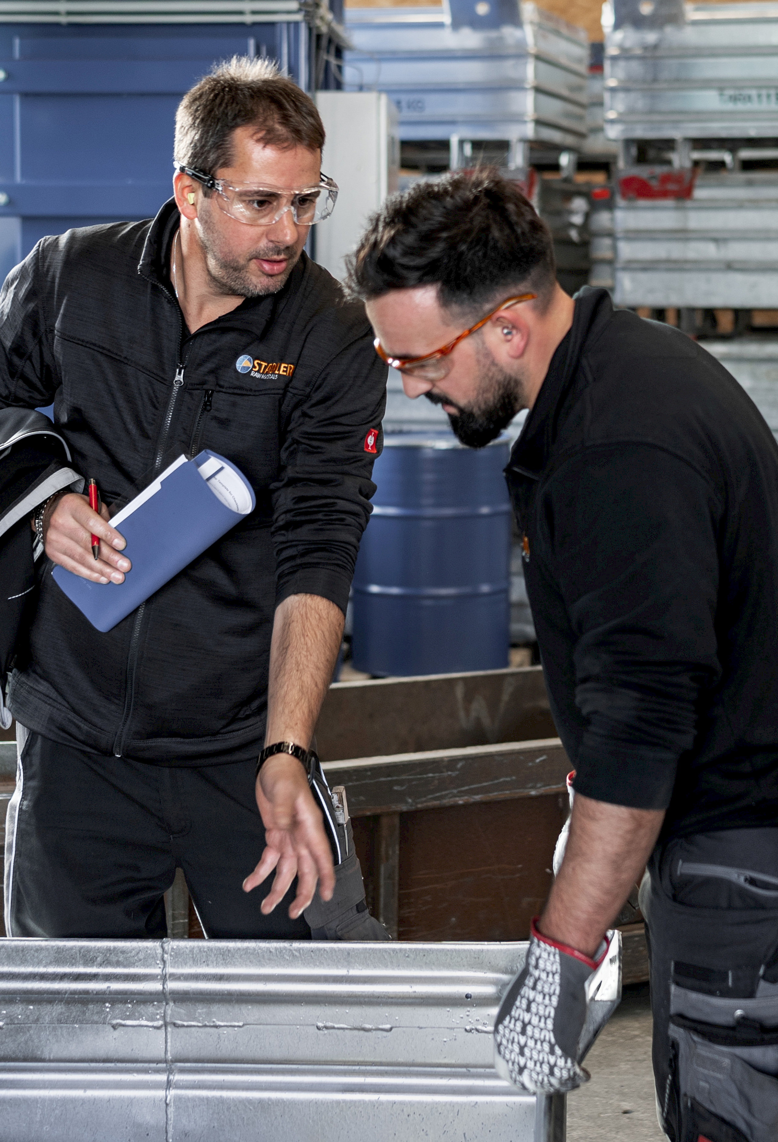 Zwei Männer mit Schutzbrillen. Im Hintergrund ist eine Lagerhalle zu sehen.