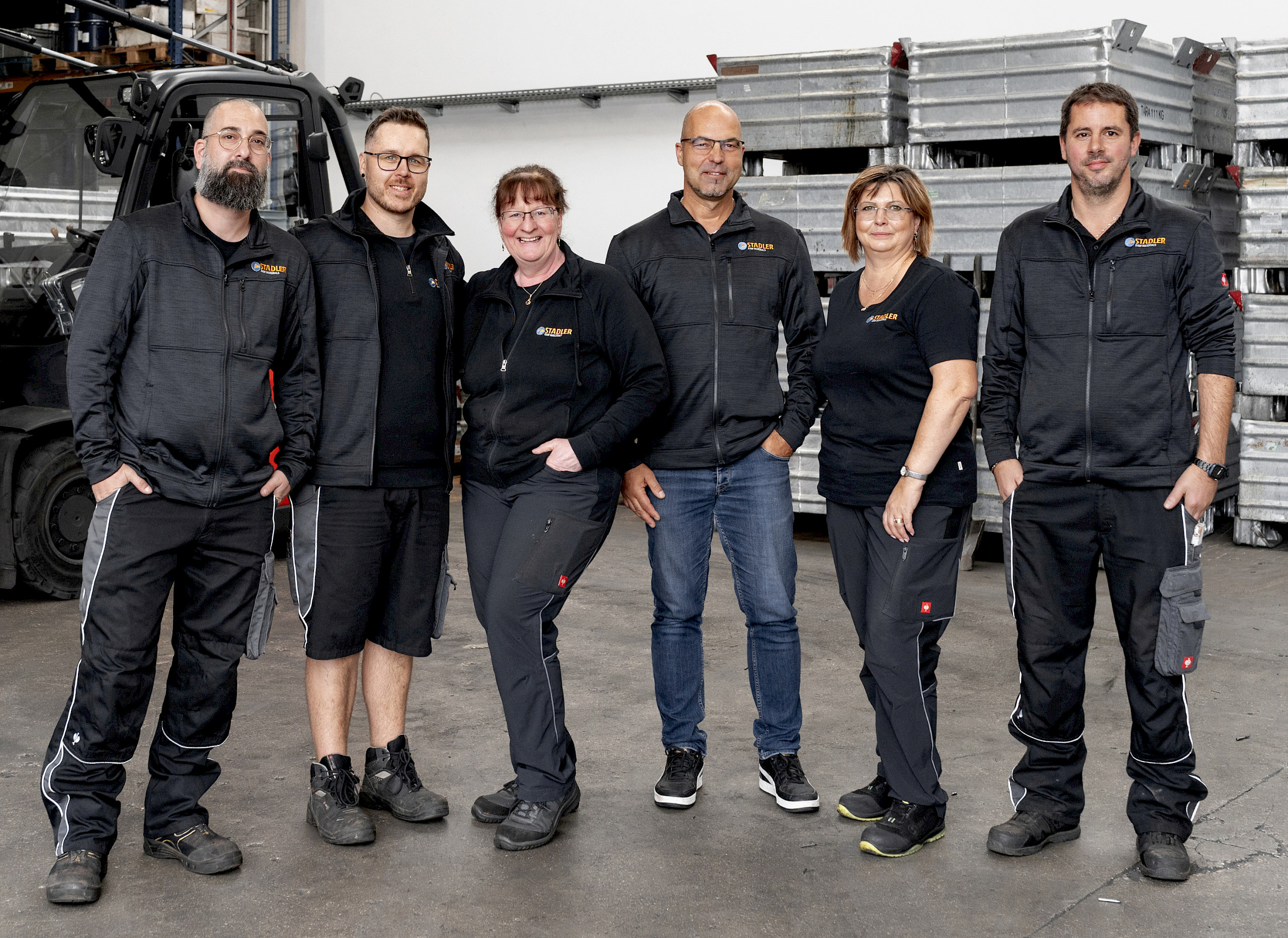 Das Team von Stadler in Form von sechs Personen in Arbeitskleidung steht in einer Lagerhalle. Im Hintergrund sind gestapelte Gegenstände sichtbar.